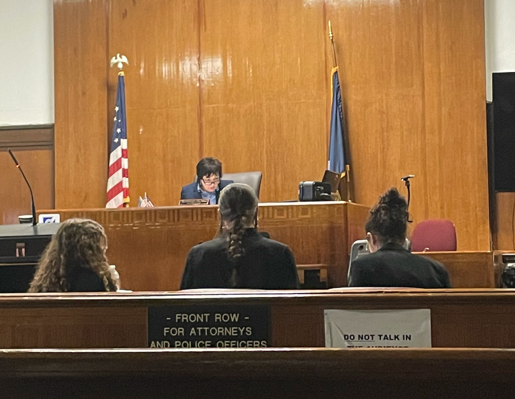 Three Student attorneys sit in a court room with their backs to the camera as they look at the judge seated on the bench.