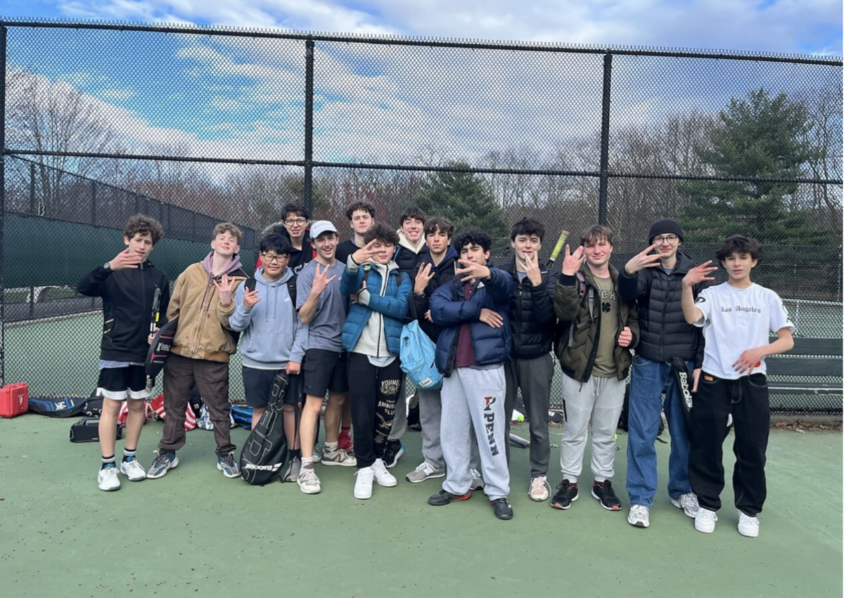 12 members of the boy's tennis team stand facing the camera on a tennis court.