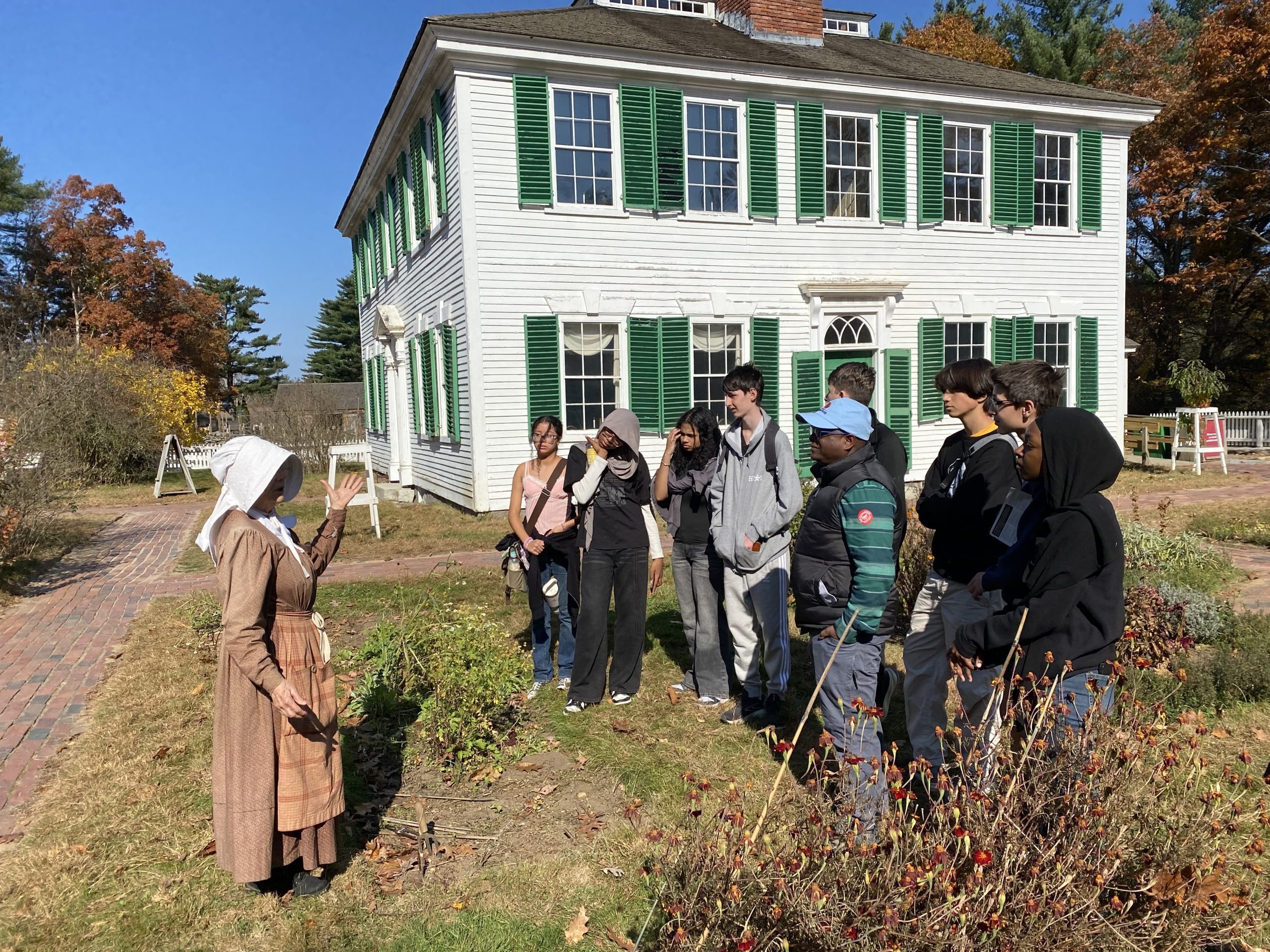 A  townsperson in 19th century dress faces 12 young people in front of a house.