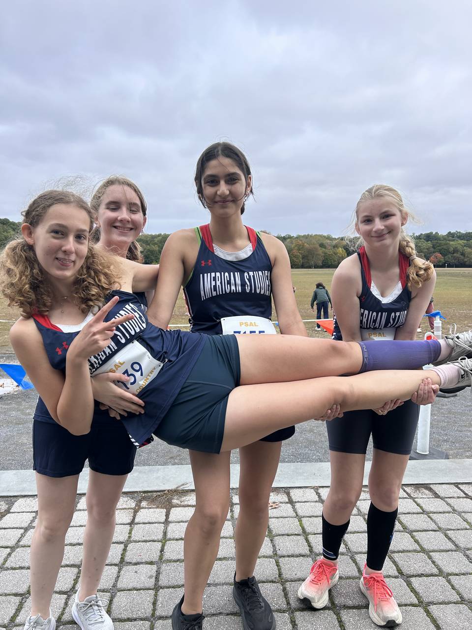 Three girls standing side by side are wearing running uniforms and they are carrying another girl