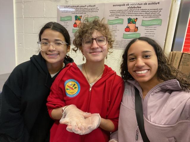 Three students stand shoulder to shoulder holding food in their hands.