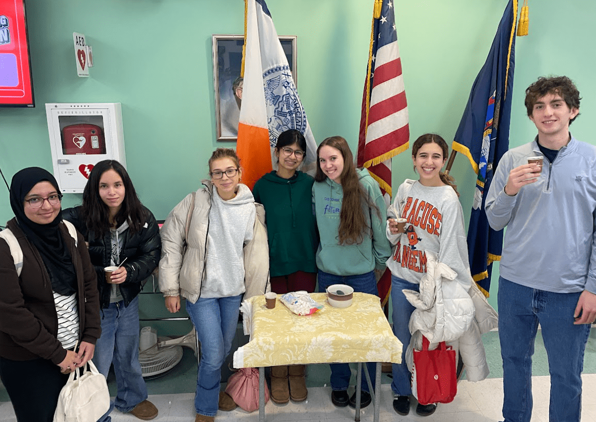 7 members of NHS stand shoulder to shoulder behind a table distributing marsmellows. 