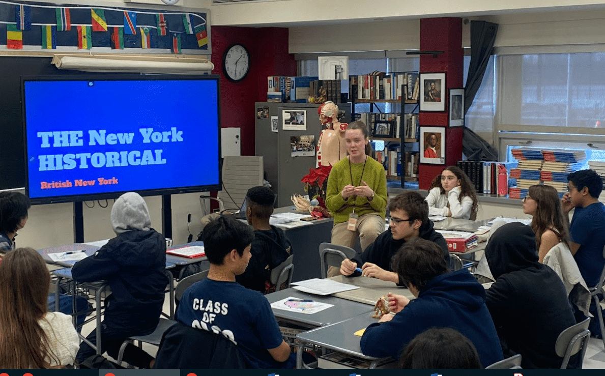 A teacher stands in the midst of sitting students explaining an artifact in her hand.