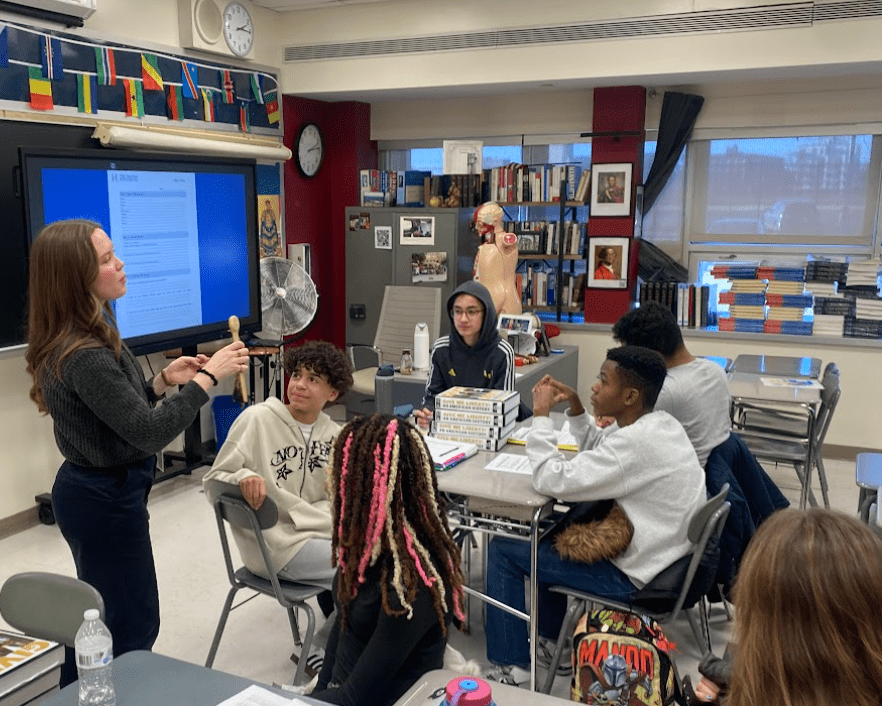 A teacher stands in the midst of sitting students explaining an artifact in her hand.