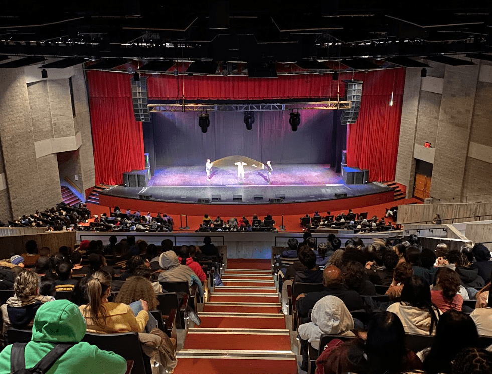 A stage within a theater is pictured with performers on stage.
