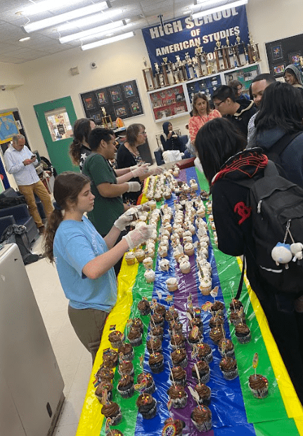 Students stand behind a long table with a rainbow color tablecloth and hundreds of cupcakes.